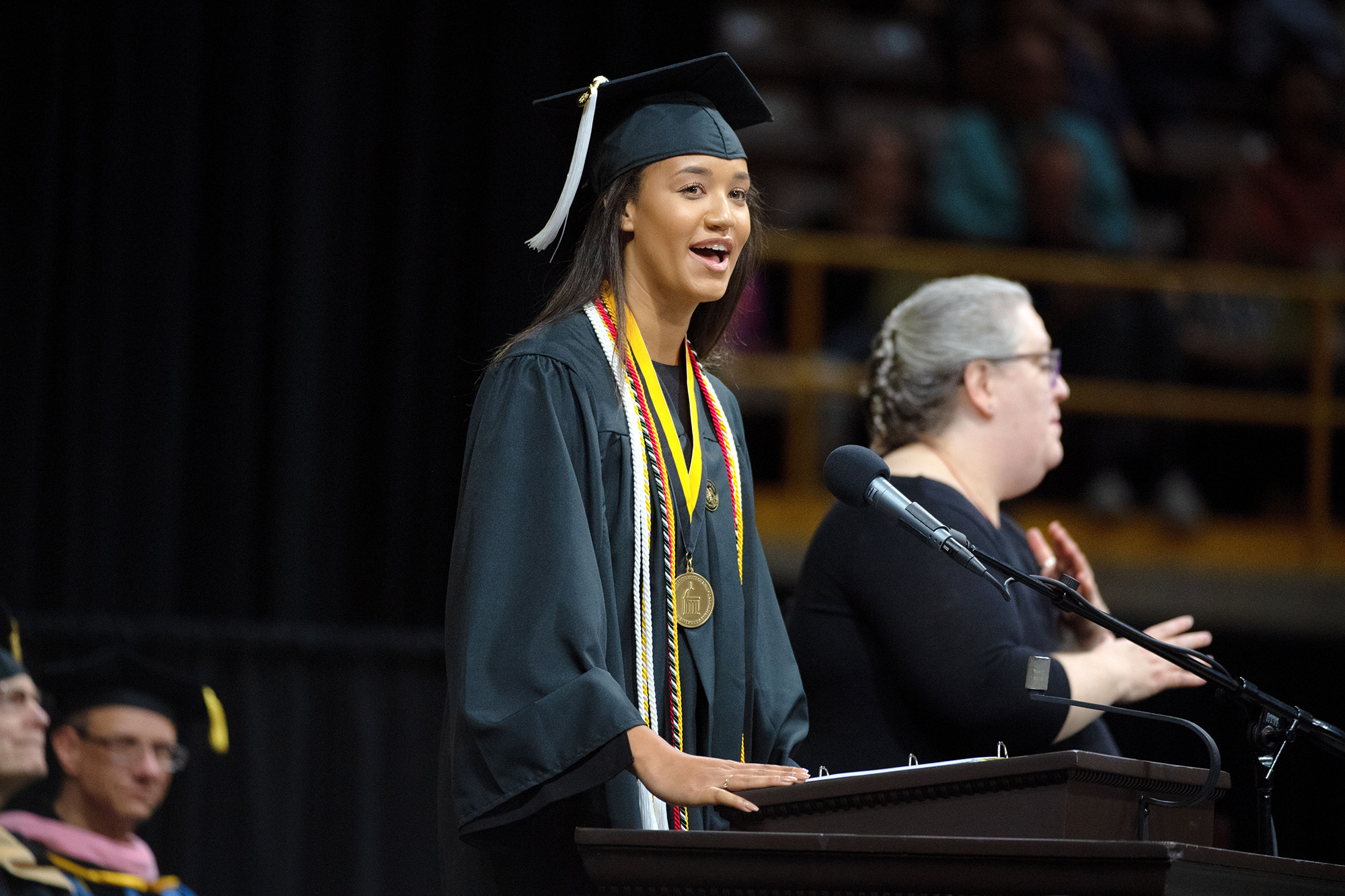 Kimmi Chex, in cap and gown, stands at a podium delivering a commencement speech.