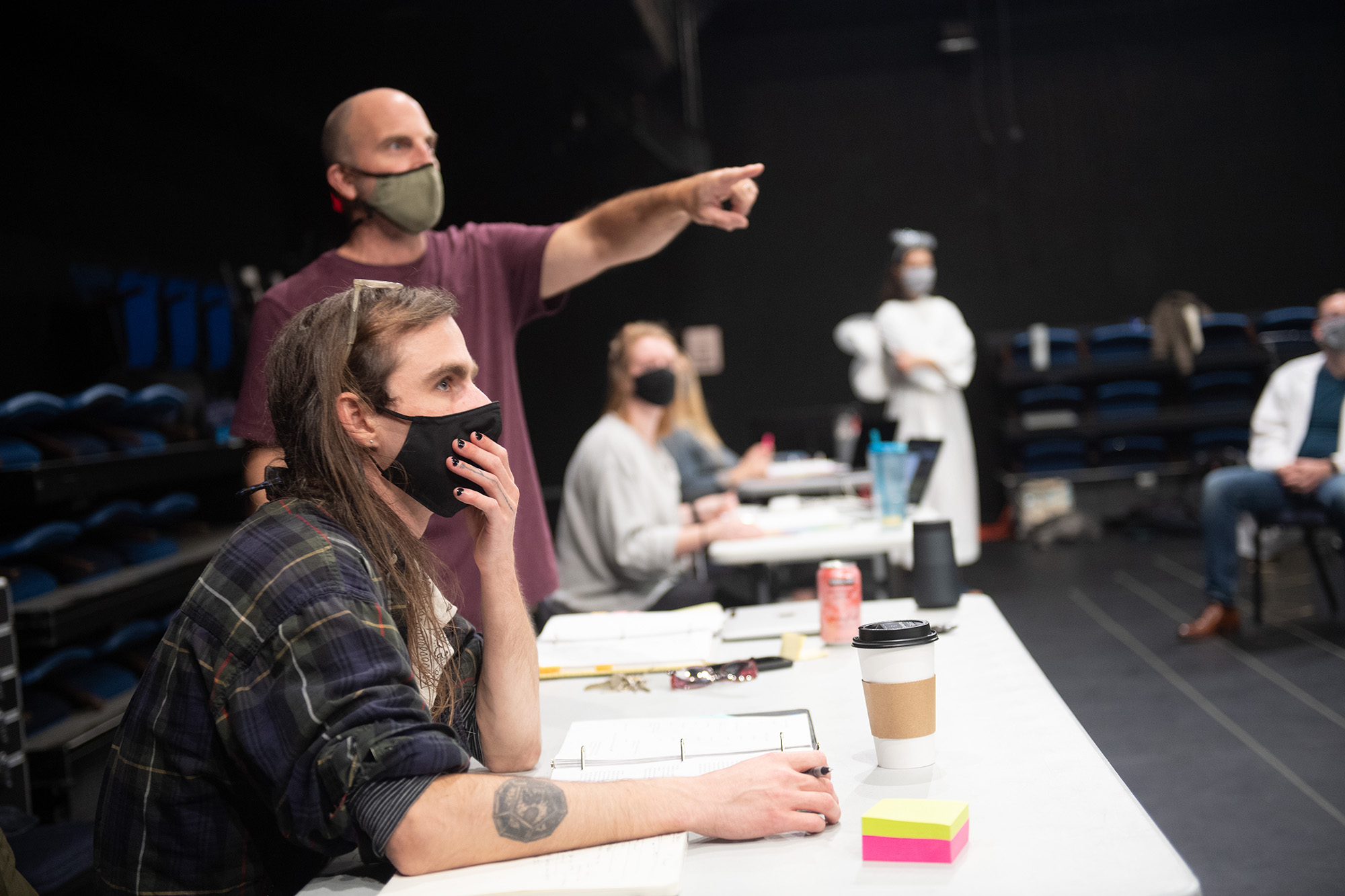 Iowa Playwrights Workshop graduate Dakota Parobek is seated on the set of their play Smile Medicine. The play's director is standing behind him and gesturing toward the cast