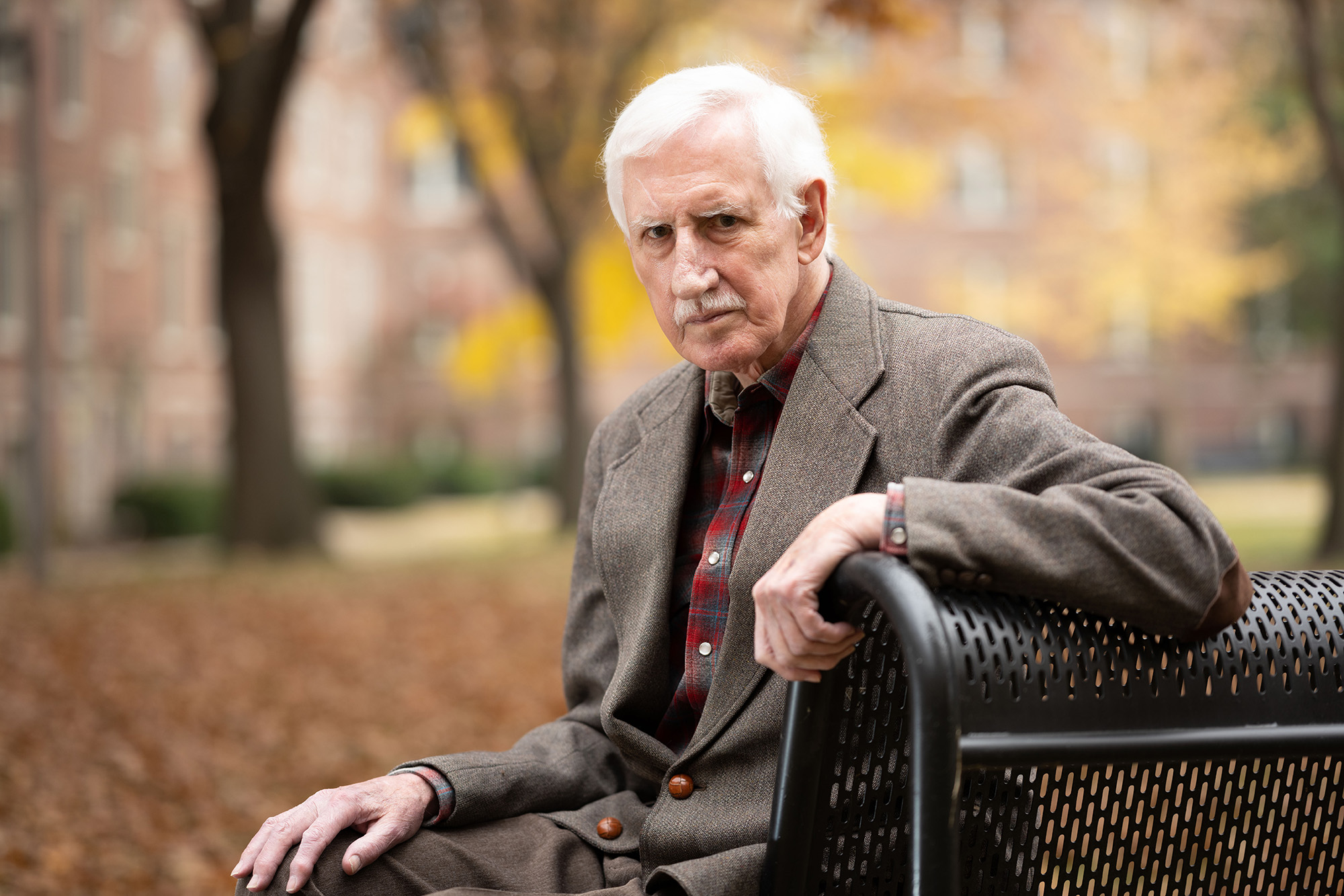 University of Iowa professor emeritus Joe Coulter sits on a bench on the University of Iowa campus with autumnal trees serving as a backdrop