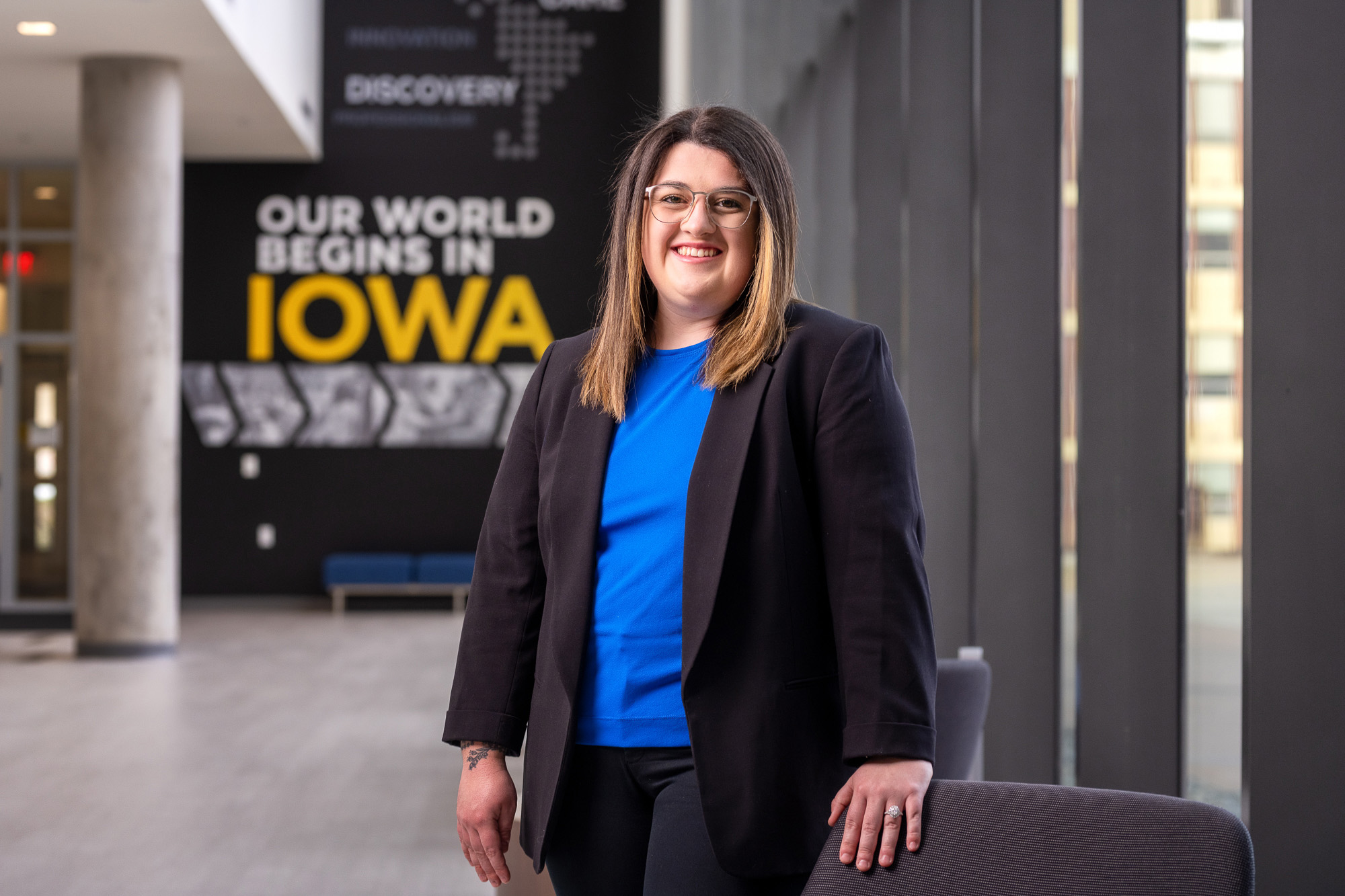 a woman standing inside the College of Pharmacy Building, her hand on a chair, with a sign that says "our world begins in Iowa" in the background