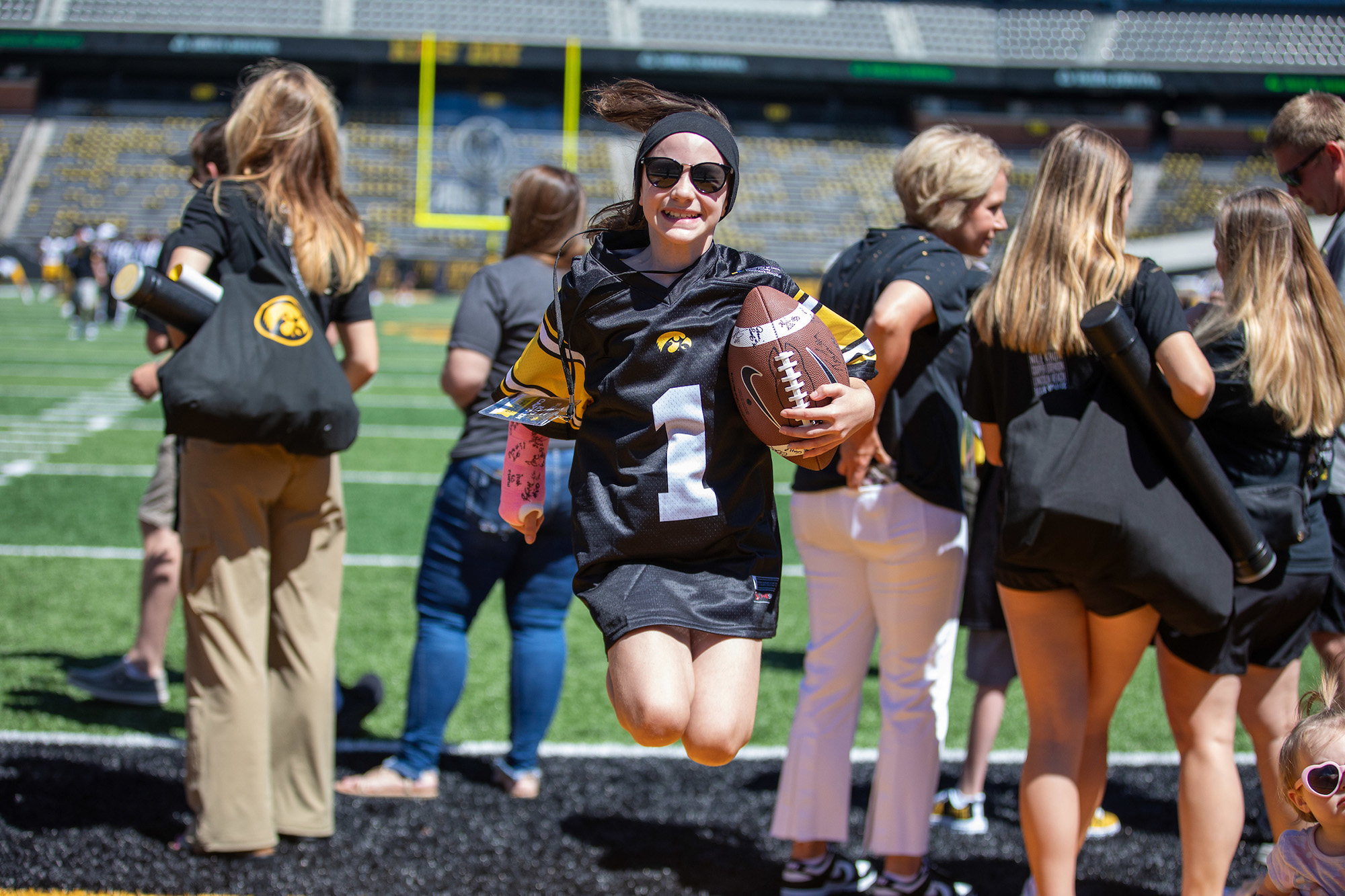 Kid Captain Gracelyn Springer jumping, wearing a Kid Captain jersey and holding a football, at Kinnick Stadium in Iowa City