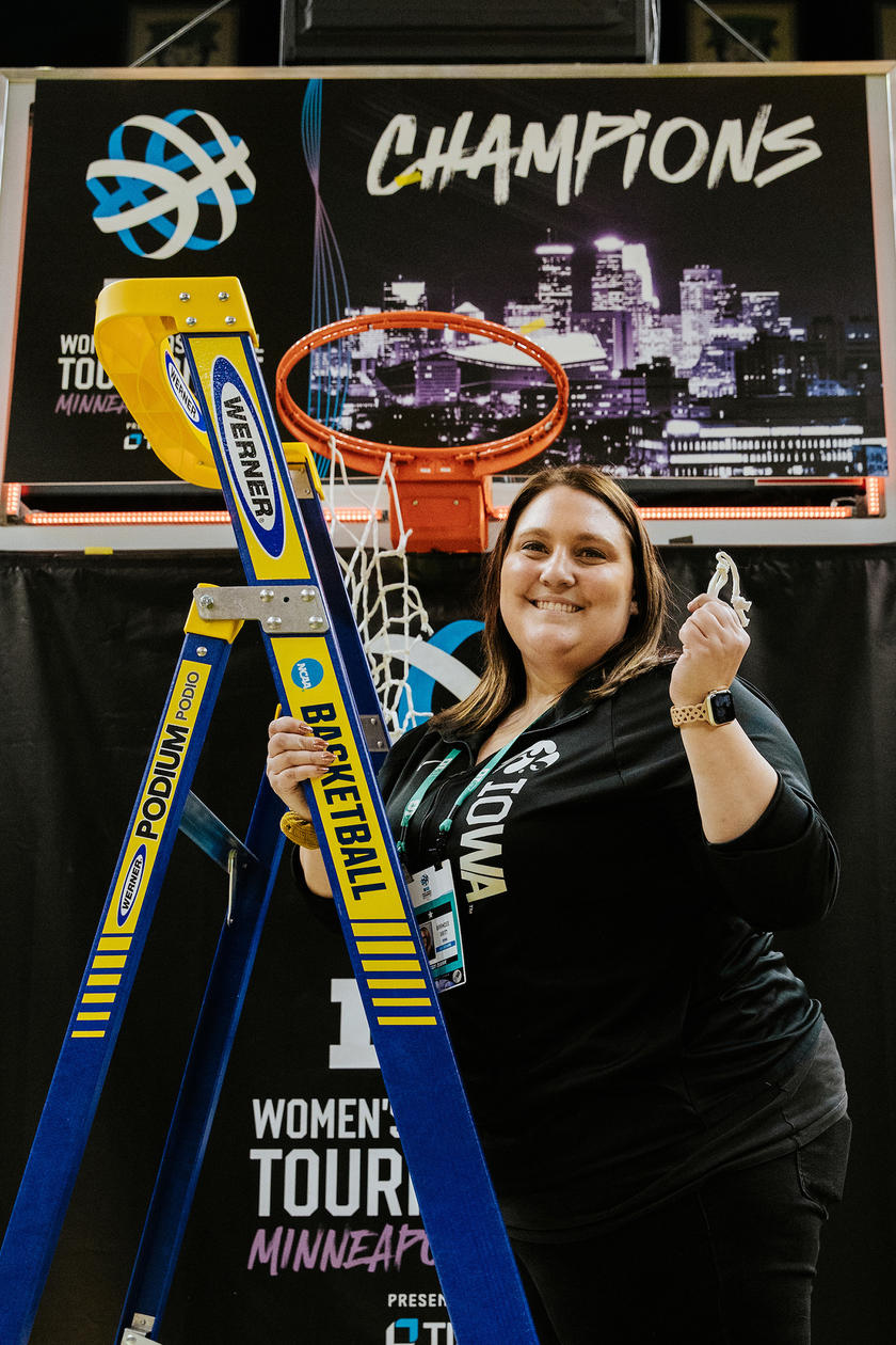 A woman standing on a ladder while smiling and cutting down the net of a basketball hoop