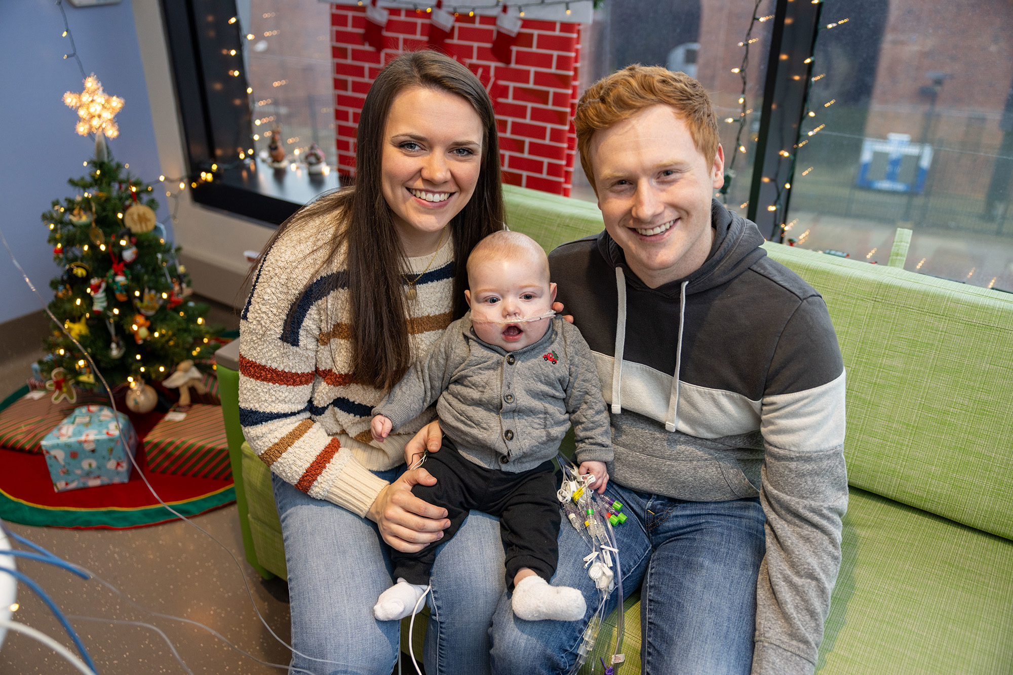 two young parents hold their infant son while they sit on a couch near a small christmas tree
