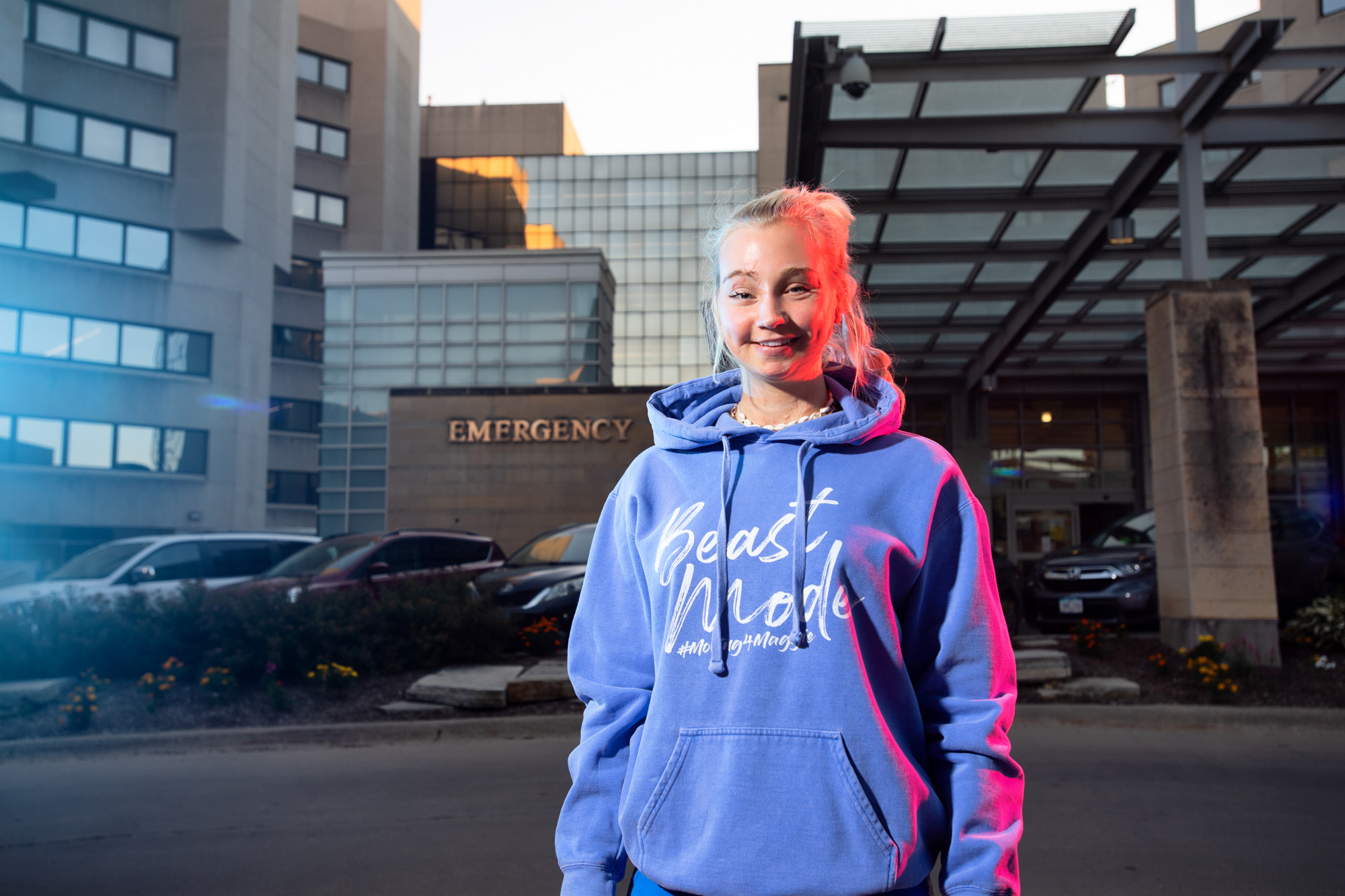 a college student wearing a "Beast Mode" sweatshirt stands in front of a hospital, with blue and red emergency lights illuminating the scene