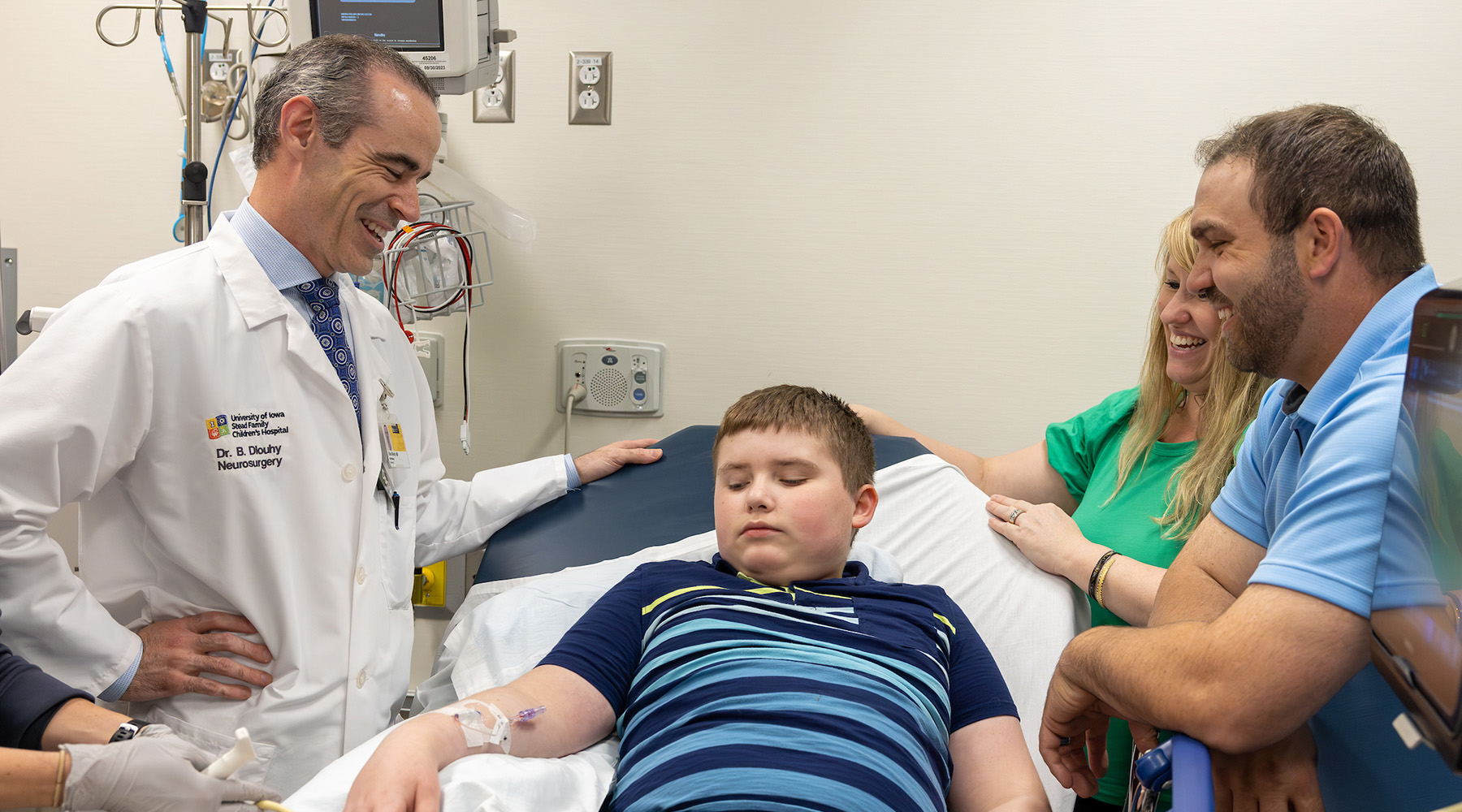 A boy laying down in a doctors office while his parents talk to a doctor