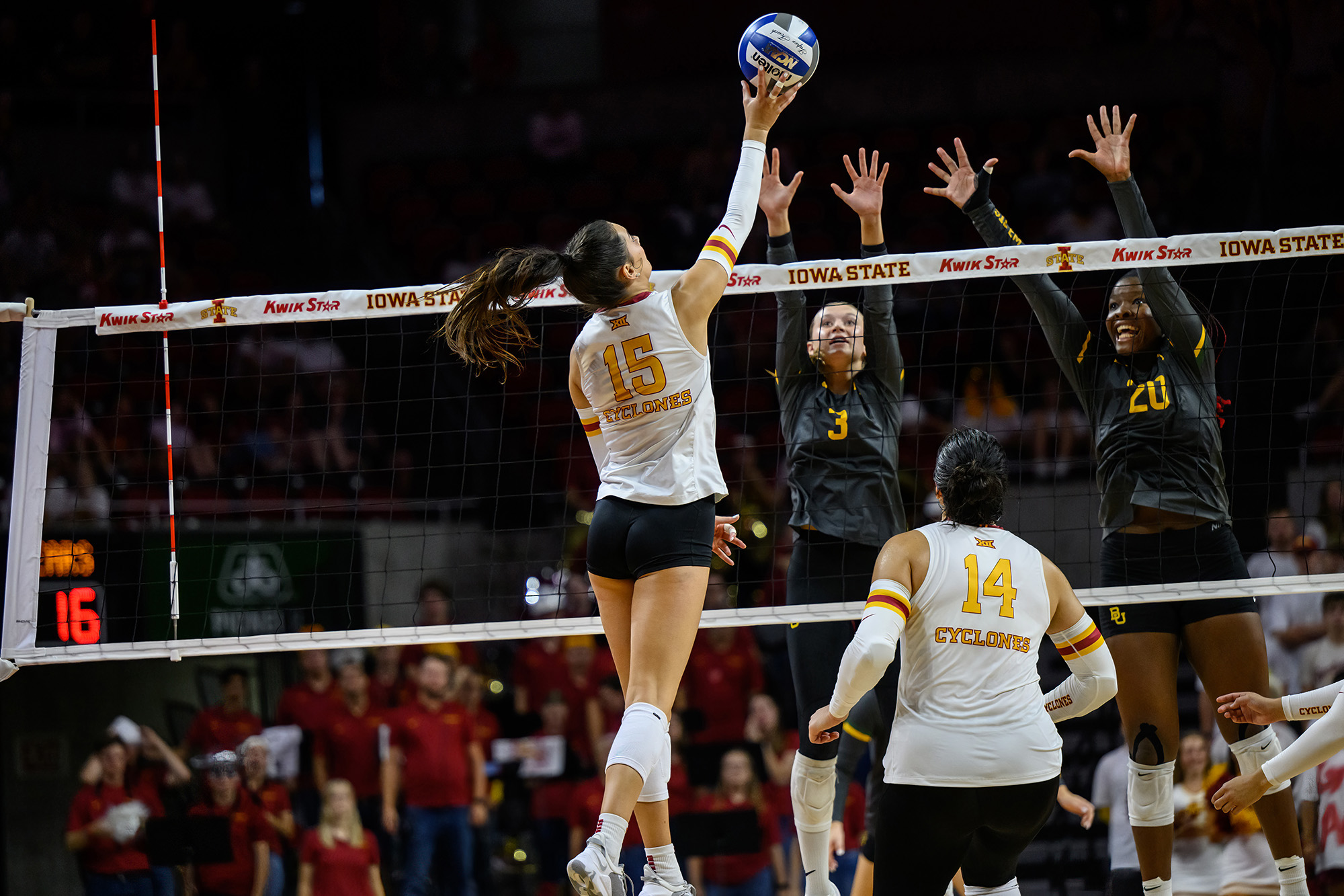 a volleyball player hits the ball as two defenders leap at the net in an attempt to block