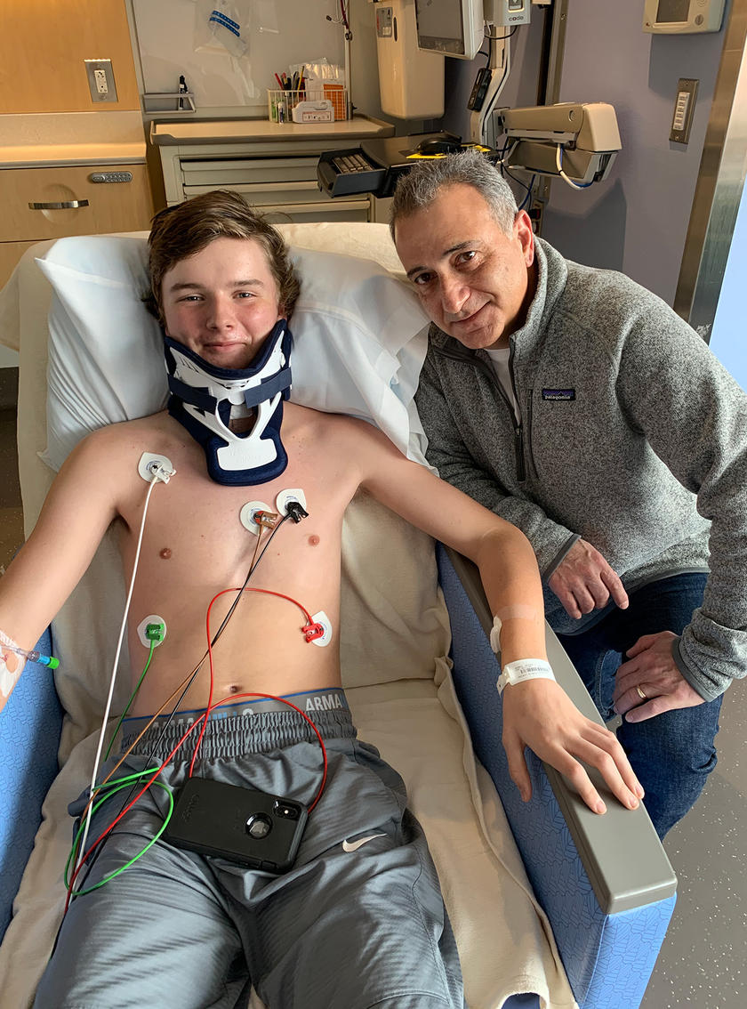 A boy laying in a hospital bed with a neck brace and wires connected to him with a surgeon kneeling next to him
