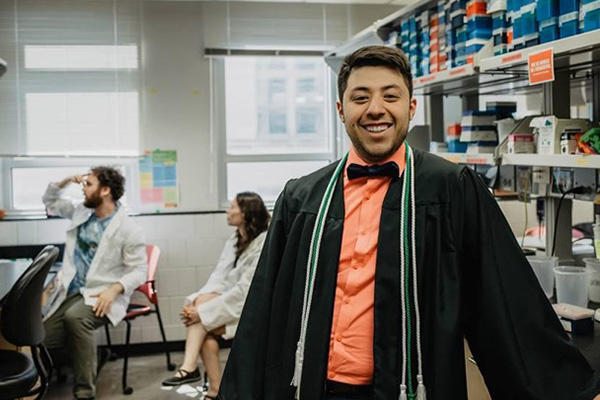A man standing in a lab smiling and wearing graduation robes