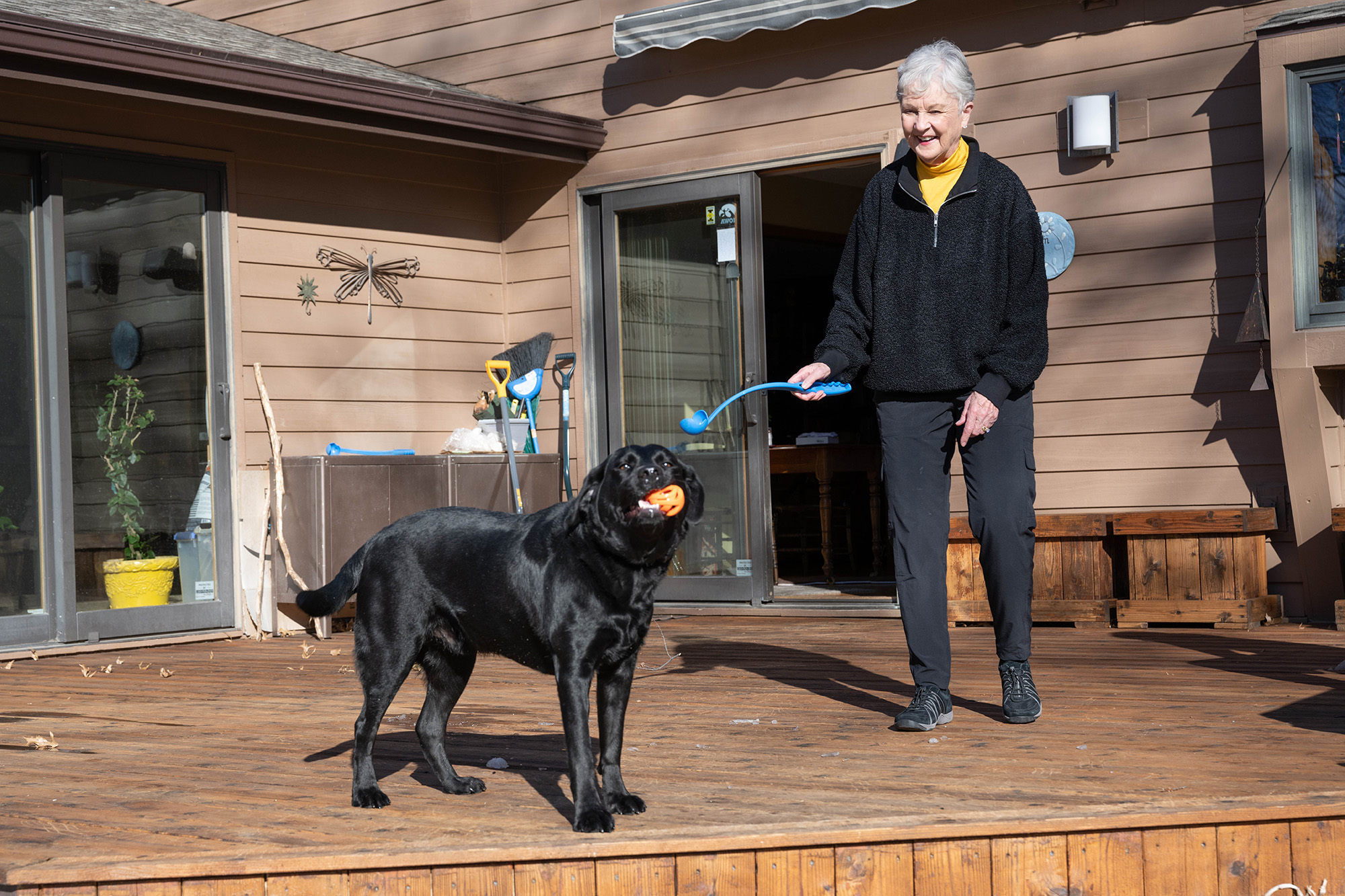 a woman and her dog on the deck at their home; the dog has a ball in its mouth, while the woman holds a tool for tossing the ball