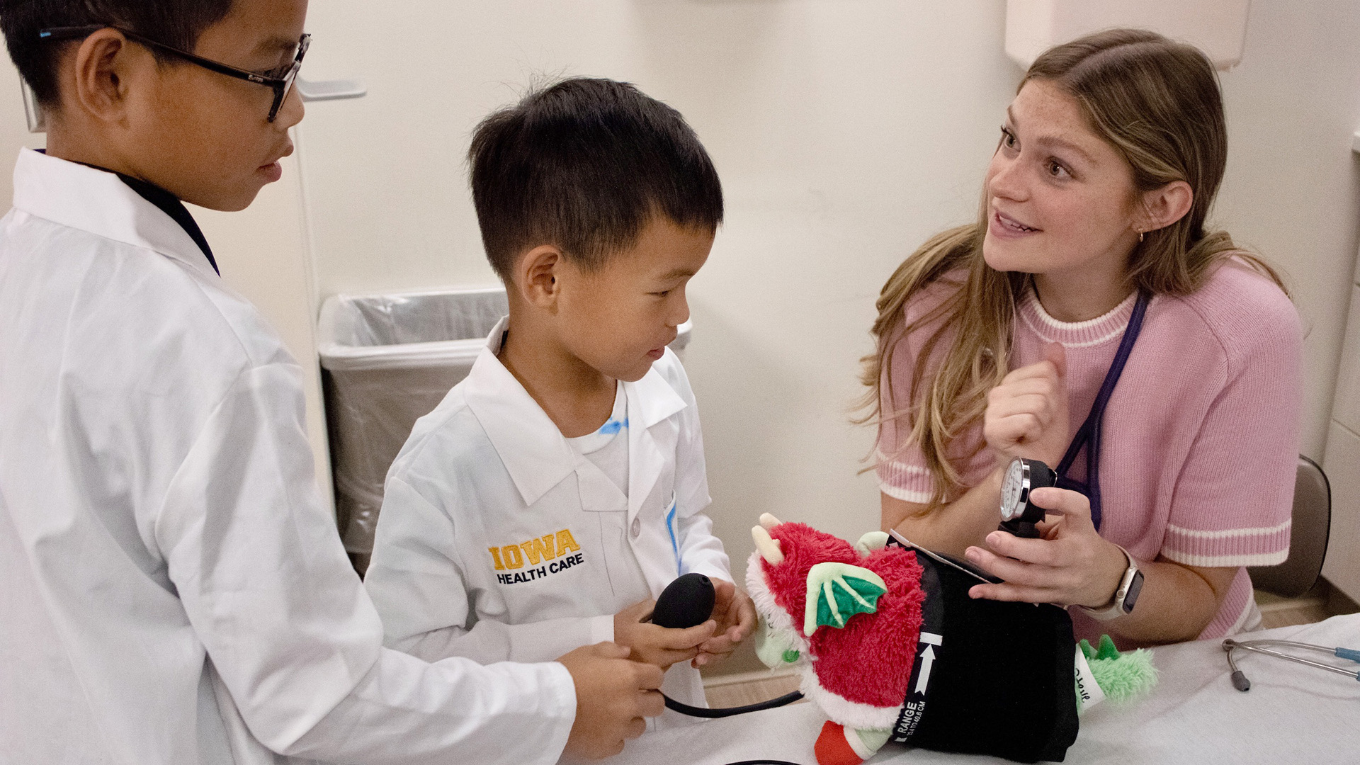 Woman in pink sweater shows a blood pressure reading on a stuffed animal to two young boys who are wearing white medical coats.