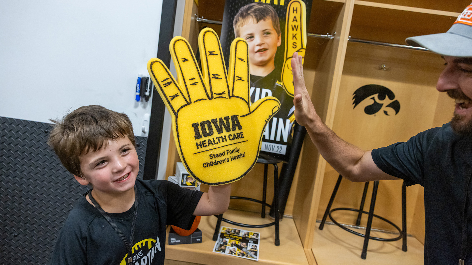 a boy wearing a large foam hand high-fives his father in a locker room