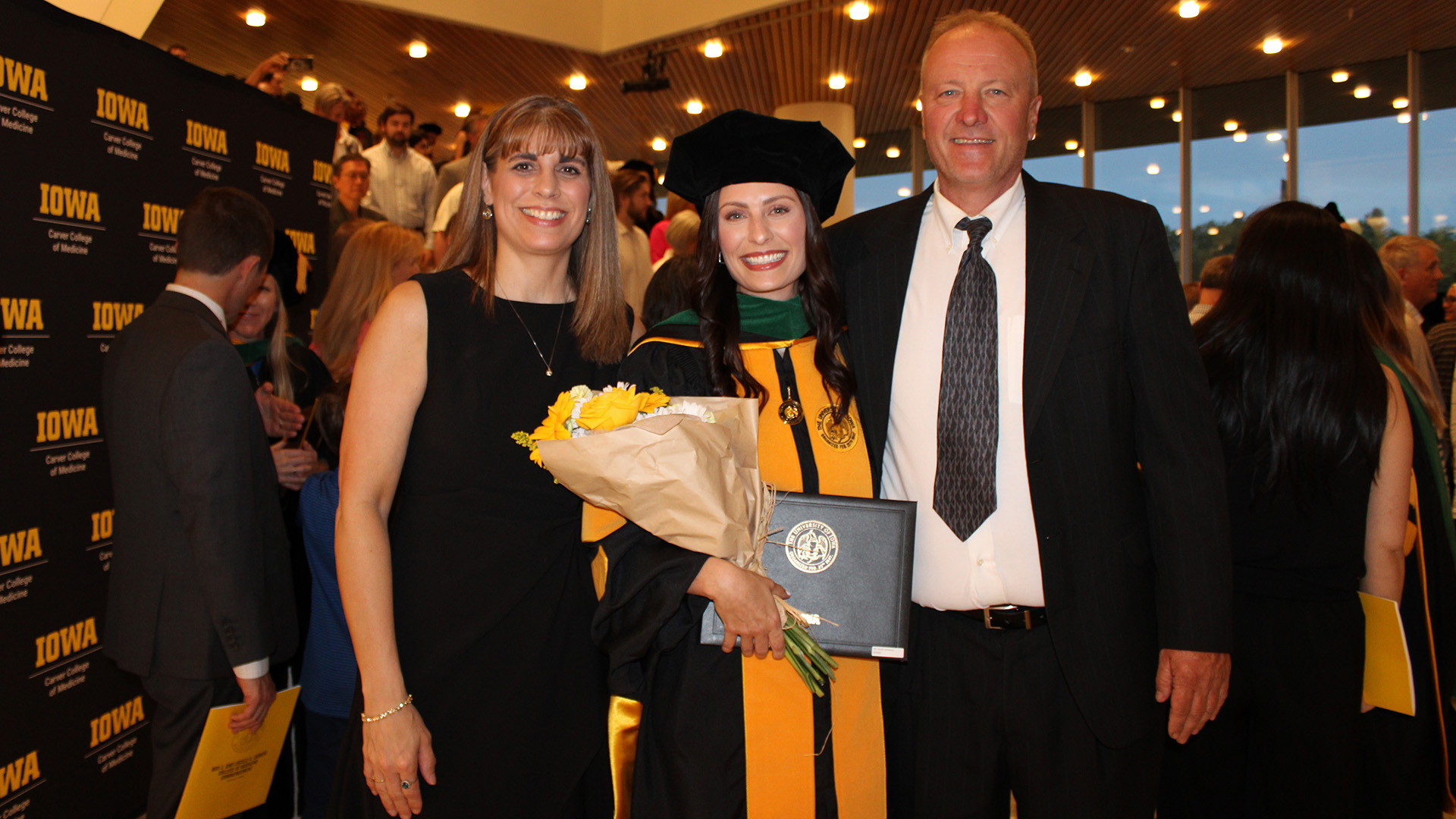 a university of iowa med school grad in cap and gown stands with her parents at a commencement ceremony