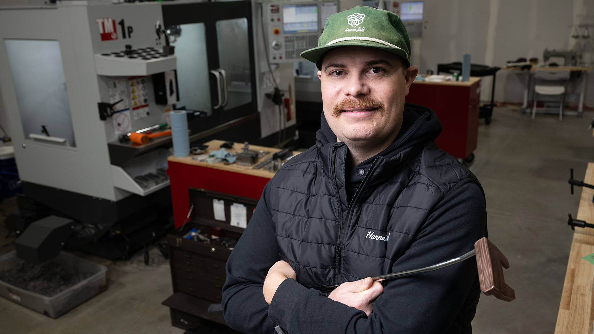 a man wearing a black Hanna Golf jacket and a green hat stands in a manufacturing facility, holding a putter