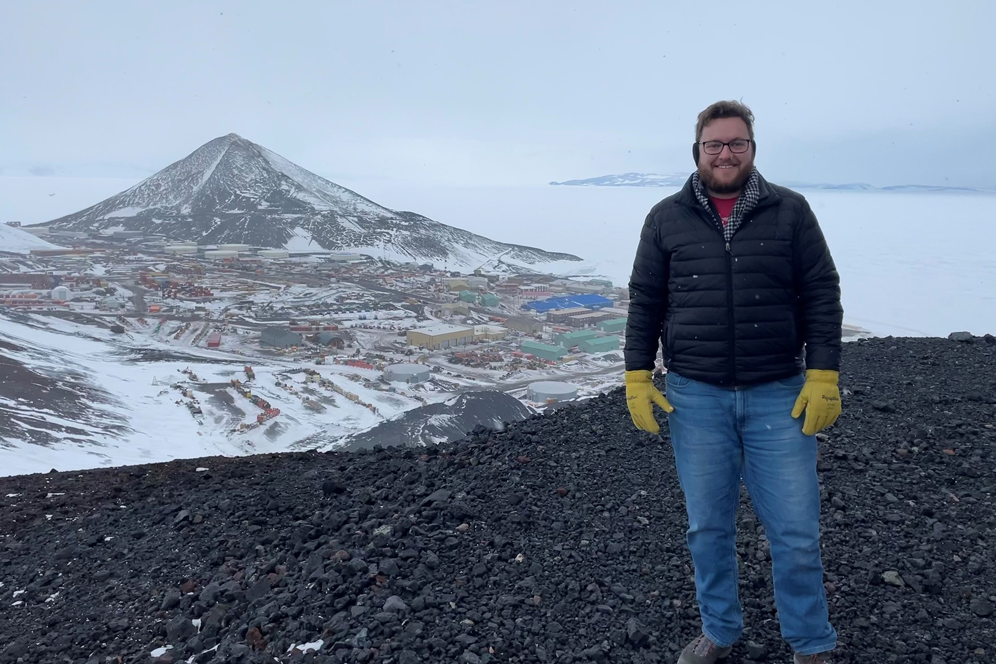 a man stands outdoors in Antarctica with buildings in the background