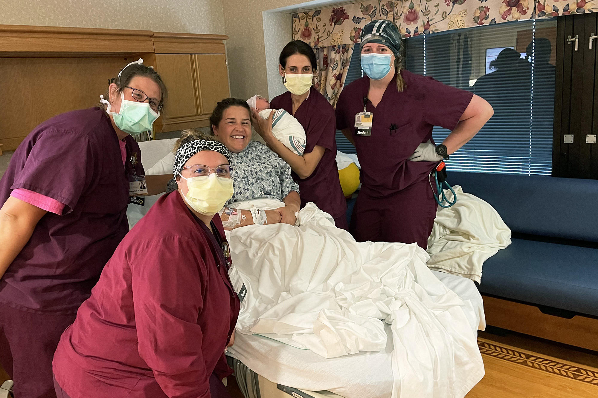 a woman in a hospital bed, surrounded by health care professionals, one of whom is holding a newborn baby
