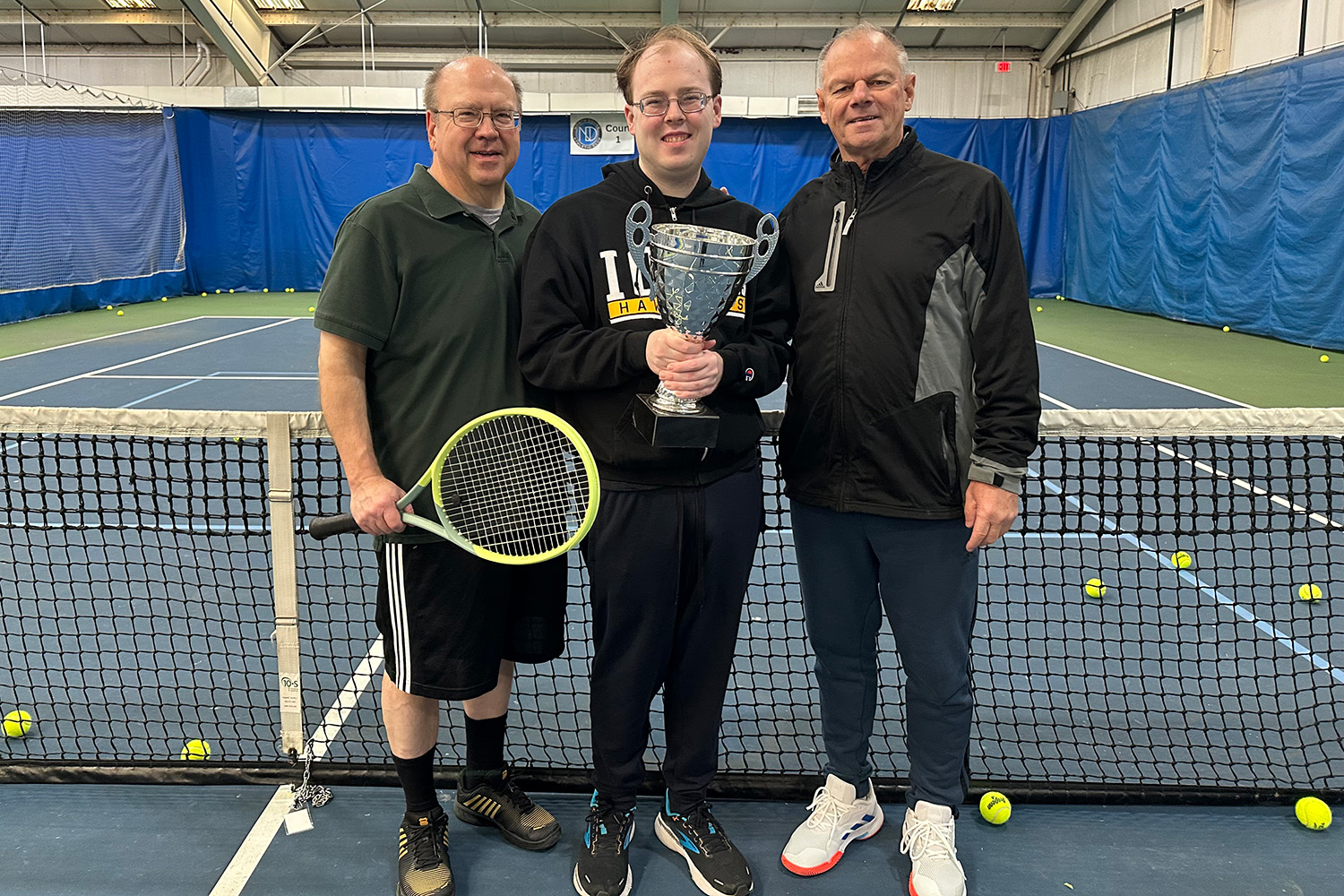 three men standing in front of the net of an indoor tennis court; the young man in the middle is holding a trophy cup