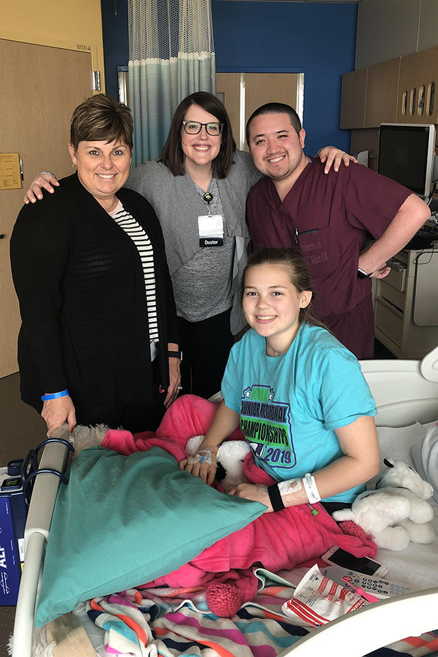 a young female patient sits on a bed flanked by her mother and two health care professionals