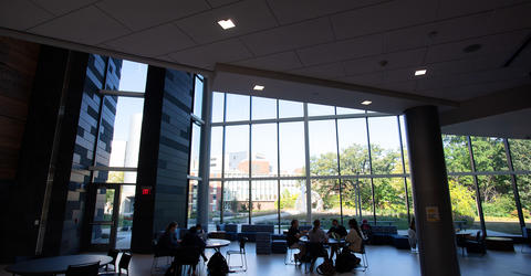 interior shot from the University of Iowa College of Pharmacy Building. People sit throughout the room; a large wall of windows shows buildings outside