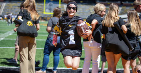 Kid Captain Gracelyn Springer jumping, wearing a Kid Captain jersey and holding a football, at Kinnick Stadium in Iowa City