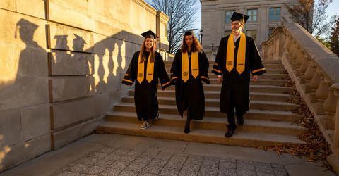 three students in cap and gown walk down steps on the university of Iowa campus