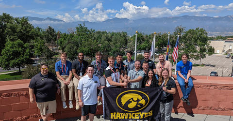 University of Iowa students and an instructor stand at the Olympic and Paralympic Training Center in Colorado, holding a Hawkeyes flag with a Tigerhawk logo on it, and a lit torch