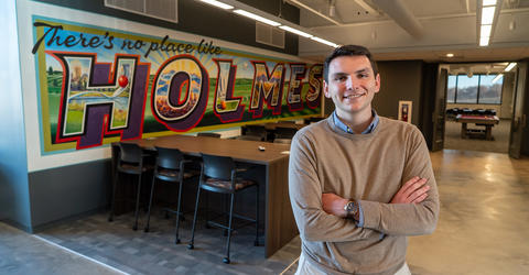 University of Iowa graduate Will Corbin standing near a wall that says There's No Place Like Holmes, which is a play on words involving his employer