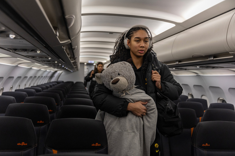 Hannah Stuelke walking down the aisle of an airplane, carrying a large stuffed animal wearing a hoodie