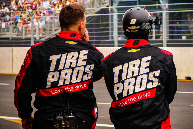 two men standing at a nascar race, wearing what appears to be team jackets