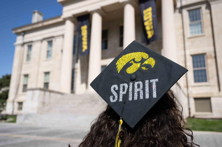 a University of Iowa student standing by Old Capitol wearing a gown and a cap with the tiger hawk logo and the word spirit