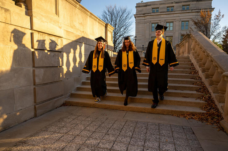 three students in cap and gown walk down steps on the university of Iowa campus