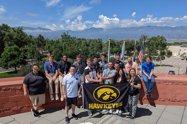 University of Iowa students and an instructor stand at the Olympic and Paralympic Training Center in Colorado, holding a Hawkeyes flag with a Tigerhawk logo on it, and a lit torch