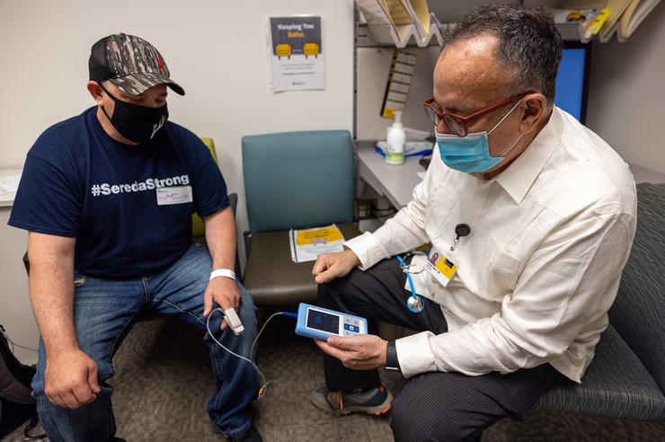 a patient and a medical professional in an exam room; the medical professional is monitoring the patient's vitals such as oxygen levels using a handheld device