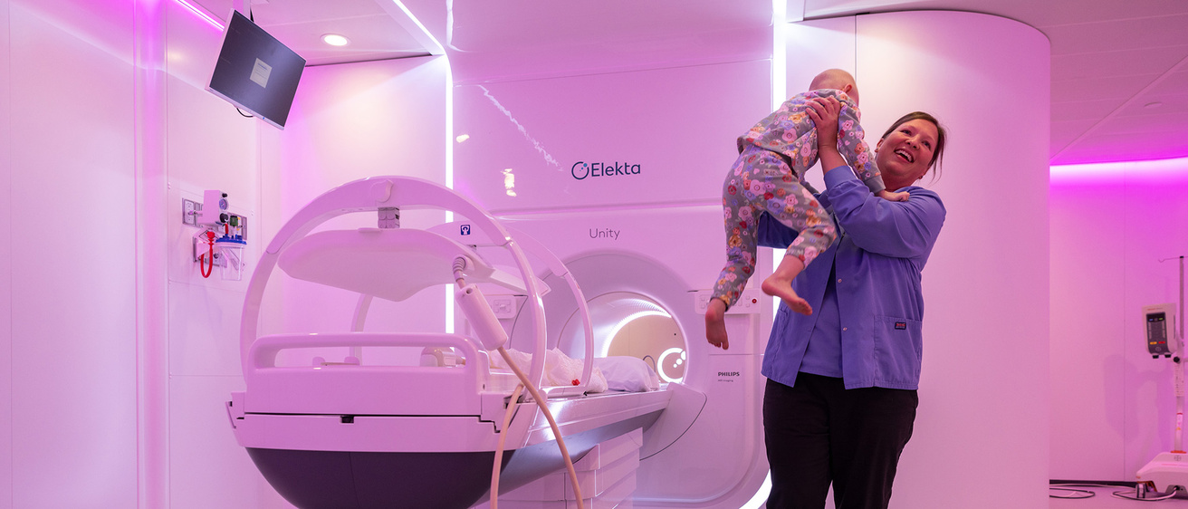 A woman holding a child and laughing while standing beside an MRI machine
