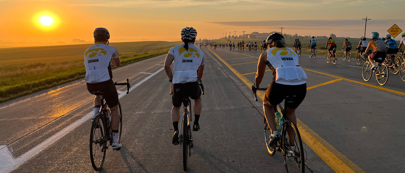 three cyclists wearing iowa tiger hawk jerseys ride along with hundreds of other RAGBRAI riders on a rural road