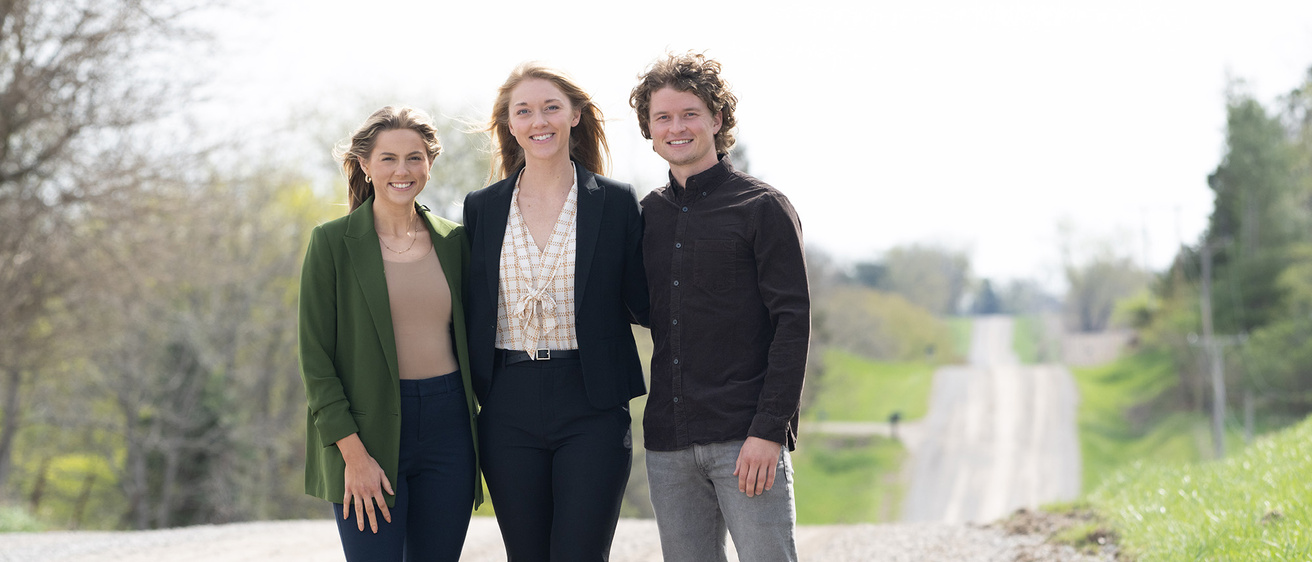 Three people stand together on a gravel road while smiling