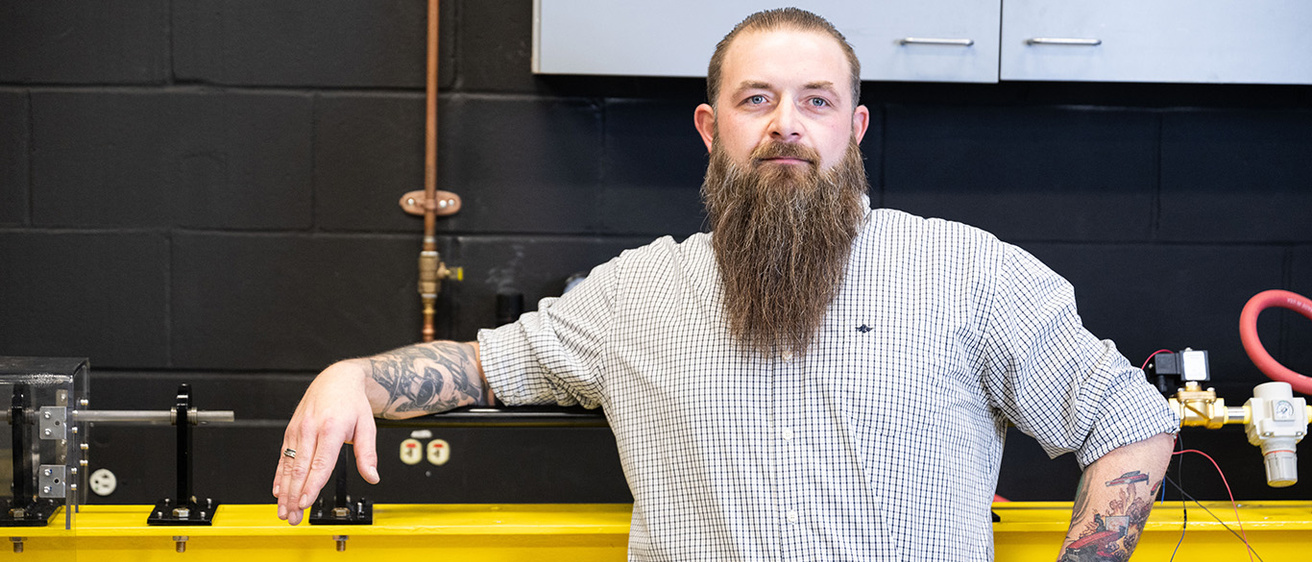 a man with a long beard stands in an engineering lab