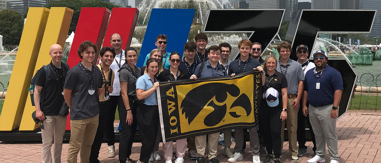 a group of people standing before a NASCAR display, holding a University of Iowa flag