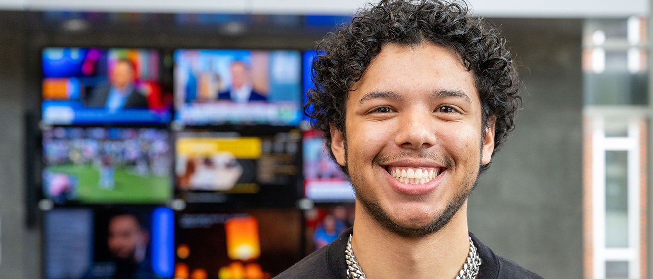 University of Iowa grad Max Willis standing in the journalism building, with tv screens in the background
