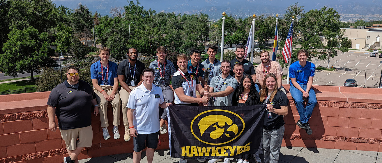 University of Iowa students and an instructor stand at the Olympic and Paralympic Training Center in Colorado, holding a Hawkeyes flag with a Tigerhawk logo on it, and a lit torch