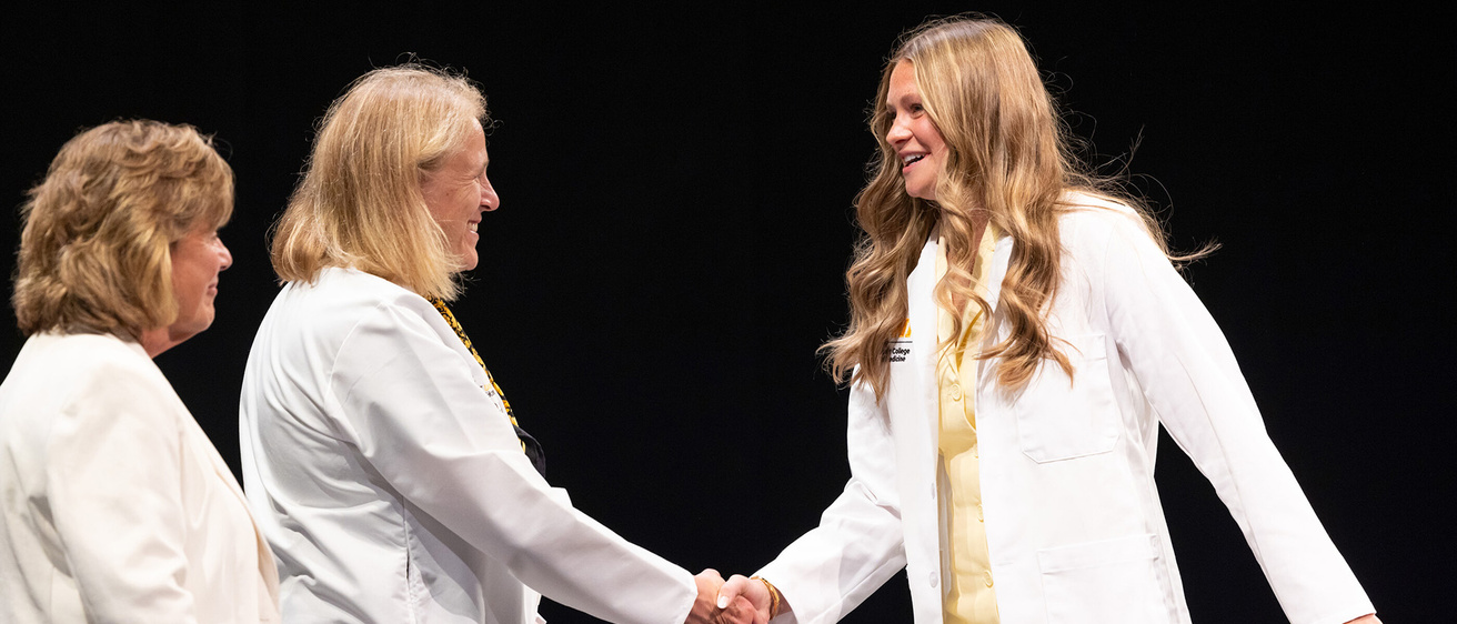 Female medical student in white medical coat shakes hands with woman who is smiling on a stage with a dark background.
