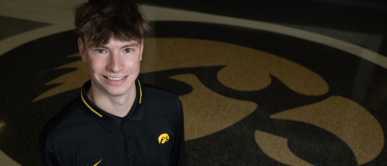 a young man wearing a hawkeye polo stands near a tiger hawk logo that is on the floor at Carver-Hawkeye Arena