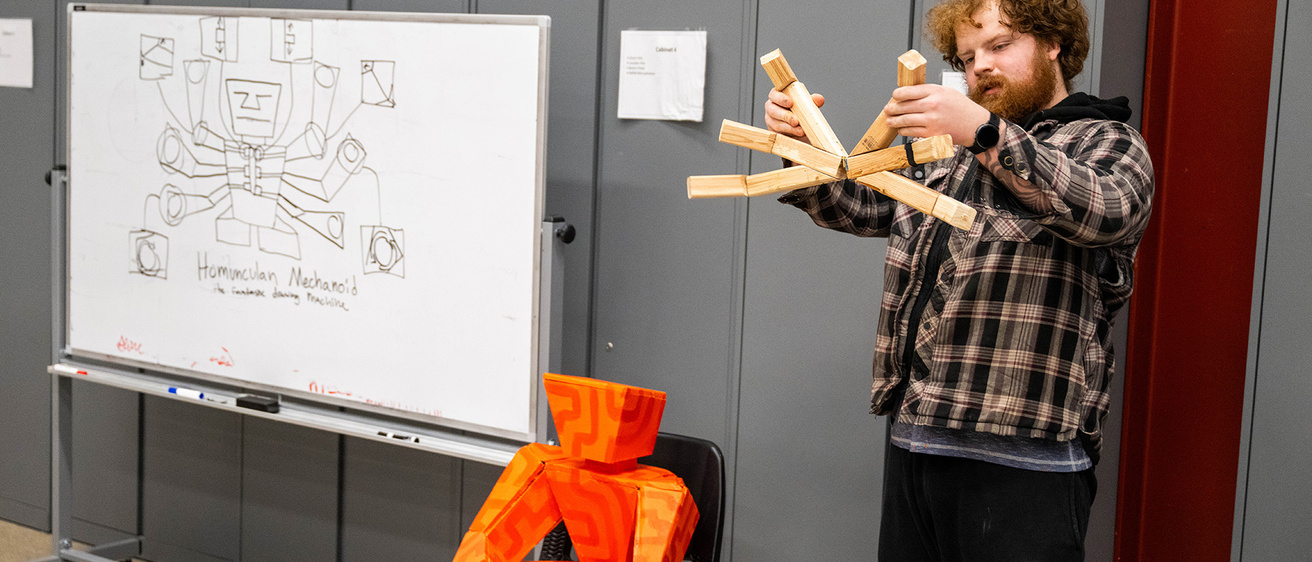 a student holds an object meant to represent arms; in the background is a sketch of a robot he hopes to create