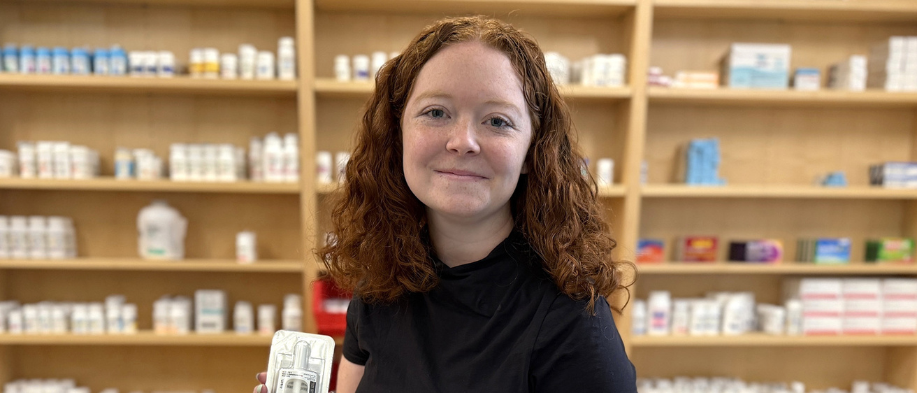 a pharmacy student holding naloxone, standing before shelves with medications