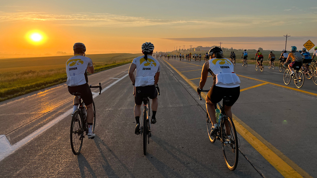 three cyclists wearing iowa tiger hawk jerseys ride along with hundreds of other RAGBRAI riders on a rural road