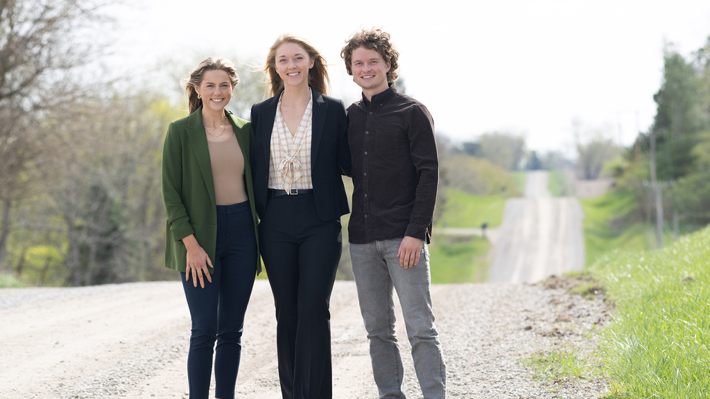 Three people stand together on a gravel road while smiling