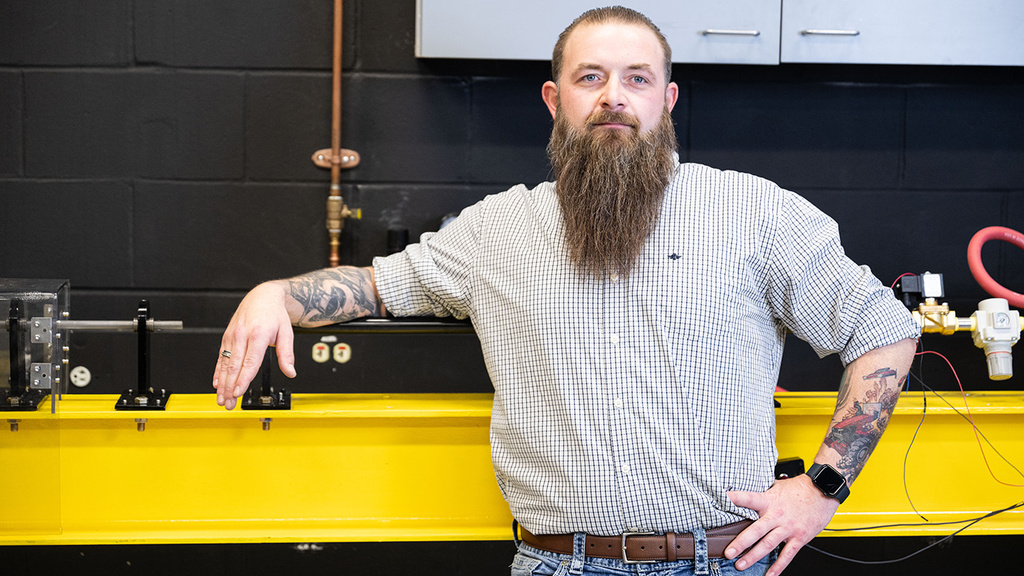 a man with a long beard stands in an engineering lab