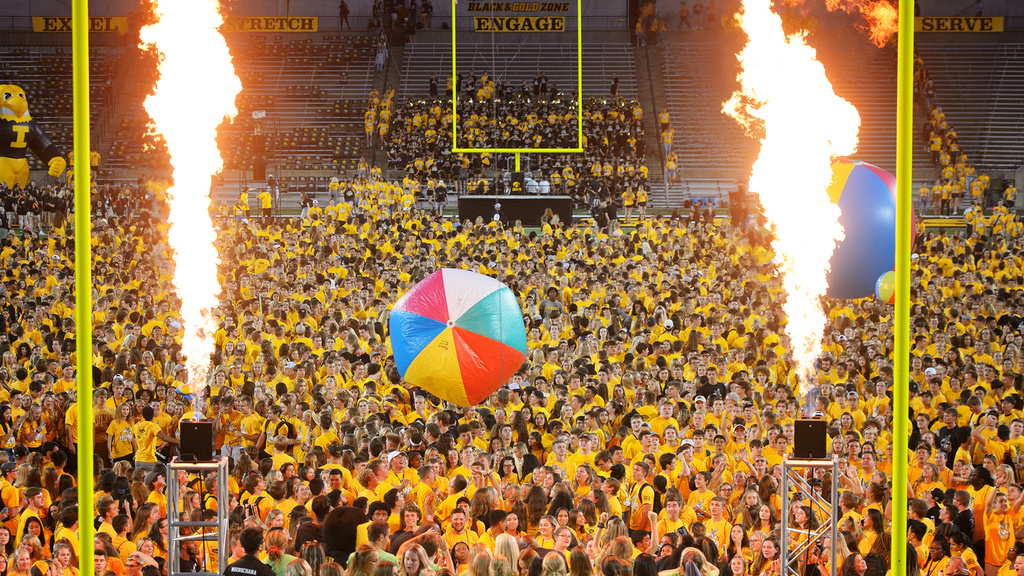 students on the field at kinnick stadium during a concert, with pyrotechnics going off and a beach ball bouncing around