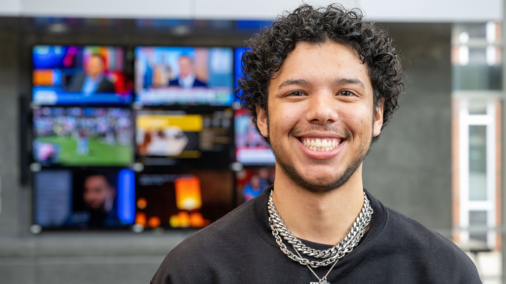 University of Iowa grad Max Willis standing in the journalism building, with tv screens in the background