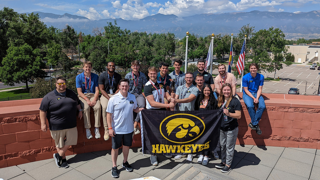University of Iowa students and an instructor stand at the Olympic and Paralympic Training Center in Colorado, holding a Hawkeyes flag with a Tigerhawk logo on it, and a lit torch