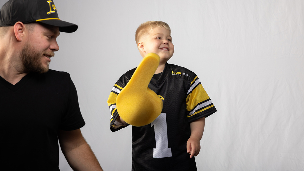 a boy wearing an Iowa football jersey holds a large foam finger while his dad watches from the side